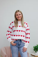 Woman wearing a white sweater with red heart patterns against a light gray background